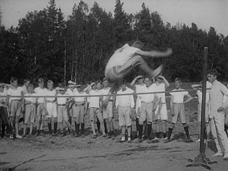 Höjdhoppare på väg över ribban på idrottstävling vid Lundsbergs skola 1913. Idrottsklädda skolpojkar i bakgrunden.