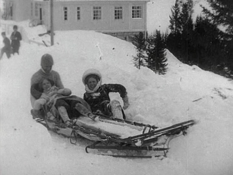 Tre vuxna på samma bobsleigh har vurpat i snön, vitt hus i bakgrunden.