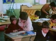 Ett par lärare hjälper unga elever som sitter i skolbänkar i ett klassrum.