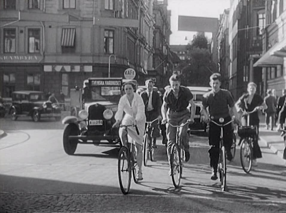 Ungdomar på cykel i stadsmiljö, bilar av äldre modell i bakgrunden.