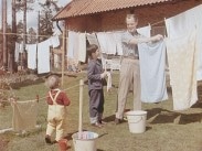 En man hänger tvätt utomhus, bredvid honom står två barn.