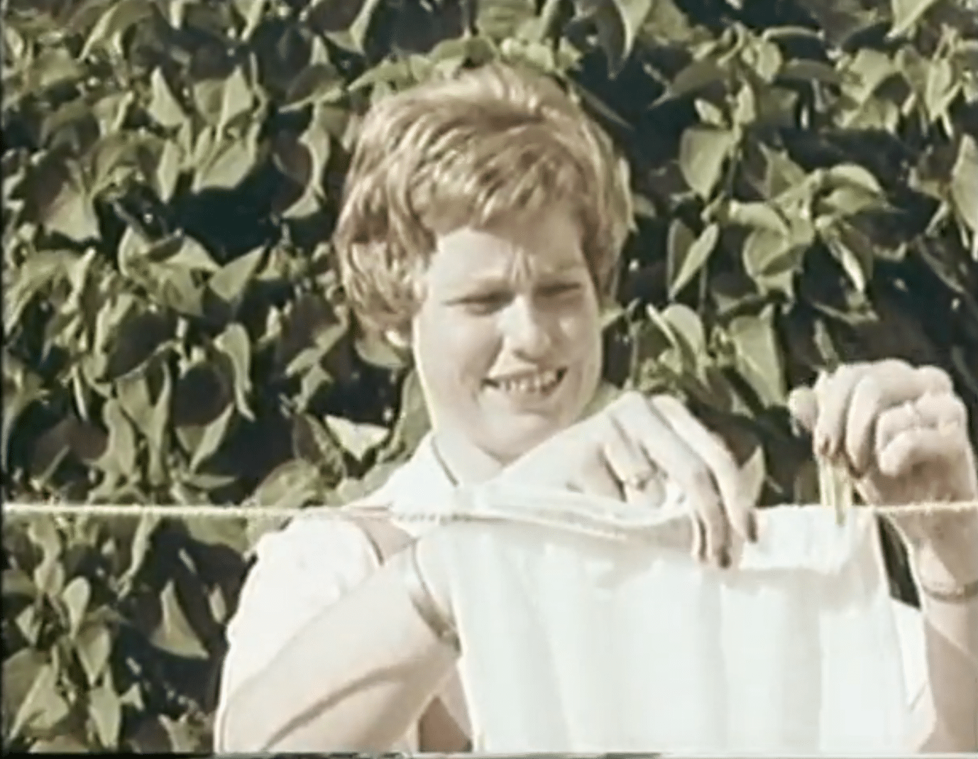 En kvinna hänger tvätt, syrenhäck i bakgrunden.