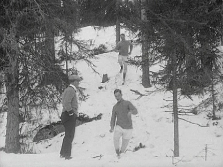 Två terränglöpare i snö springer nerför en slänt i skogen mot kameran, en man tittar på.