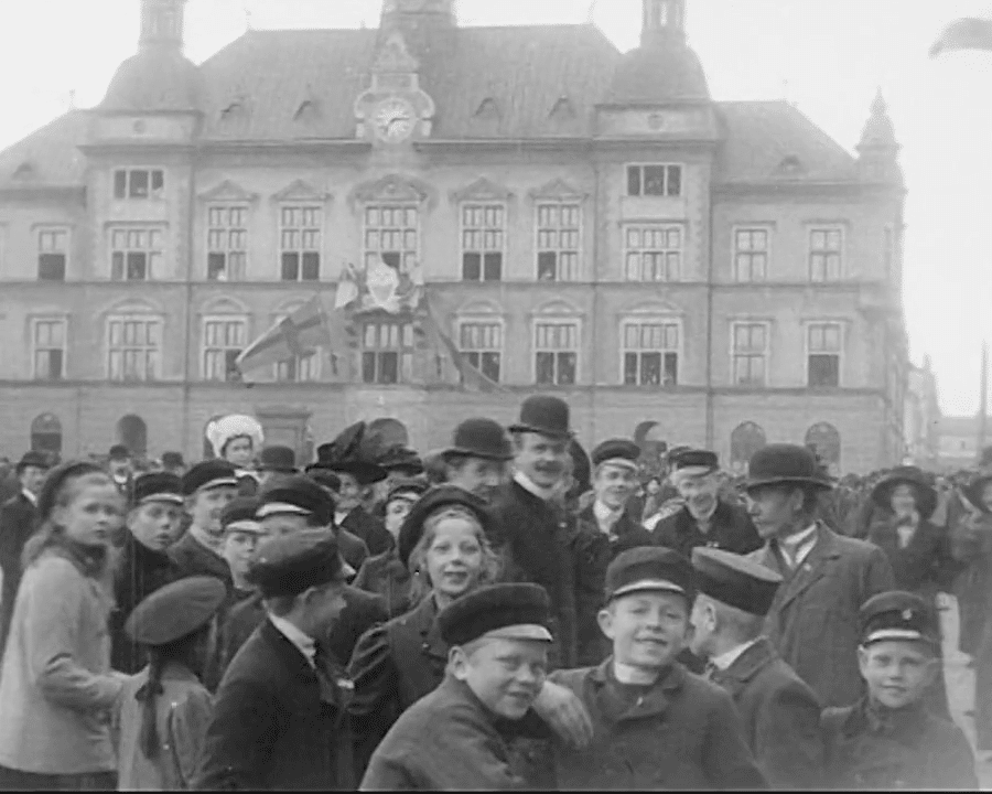 Första svenska veckan i Eskilstuna våren 1911. Folksamling på torg med byggnad i fonden, mest barn men även en del vuxna. De flesta tittar in i kameran.