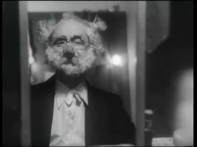 Åke Söderblom med lösnäsa och glasögon maskerad till en vithårig, skäggig gammal man ser sig själv i en spegel.
