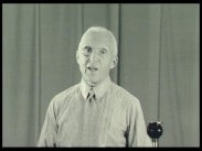 Hovstallmästaren greve Clarence von Rosen med monokel bakom en mikrofon på stativ, ridå i bakgrunden.