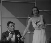 En man håller en pistol i höger hand och en gammaldags telefonmikrofon i den vänstra, bredvid står en förskräckt kvinna i vit klänning med ena handen runt mannens vänstra handled.