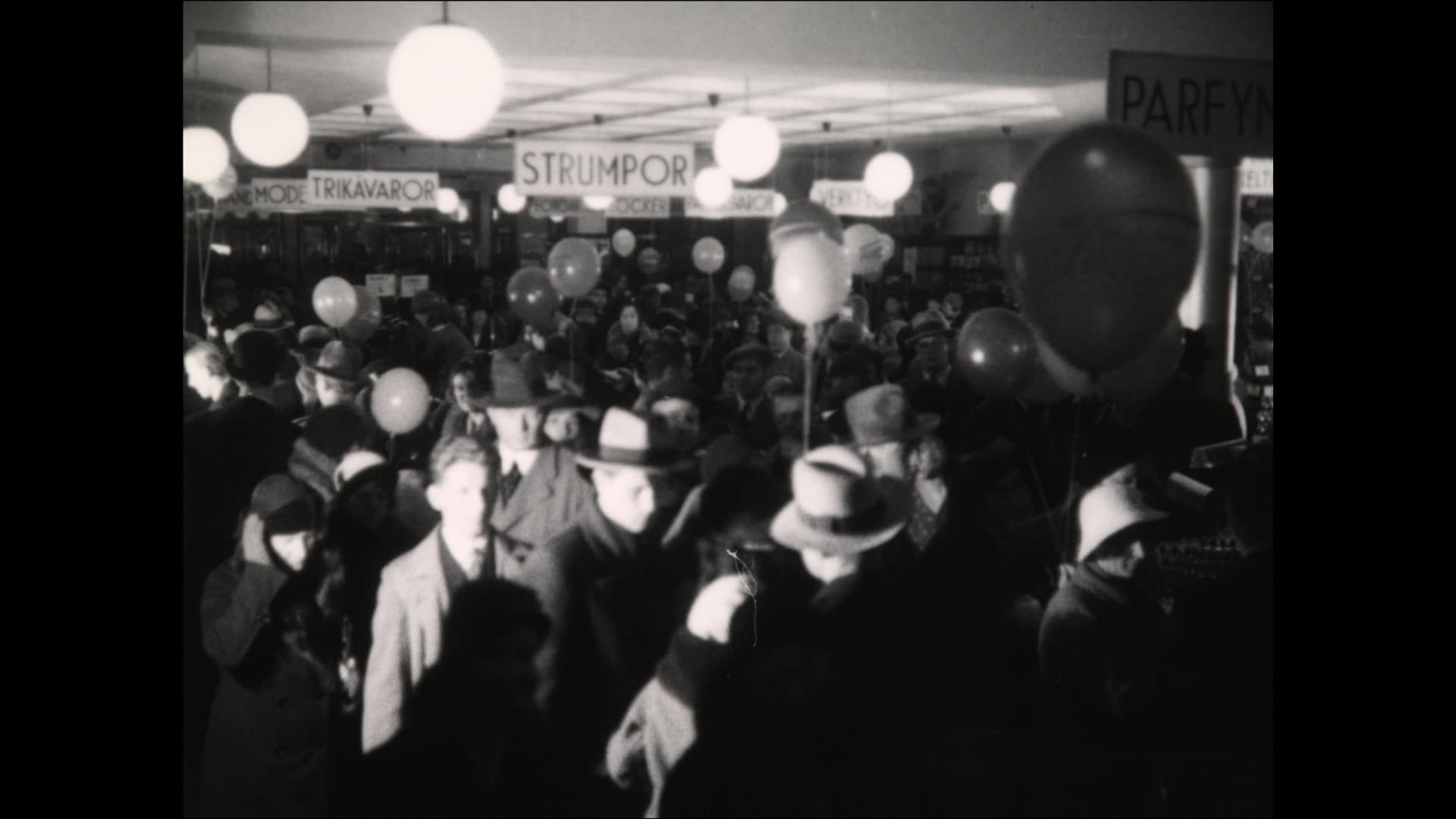 Folksamling med ballonger vid öppnandet av varuhuset Tempo i Stockholm 1932, sfäriska lampor och avlånga avdelningsskyltar hängandes från taket.