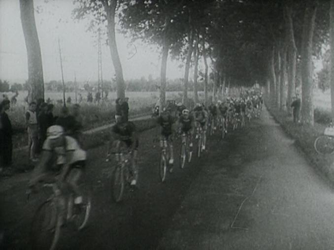 Tävlingscyklister på lång rad, träd och publik vid sidan av vägen.
