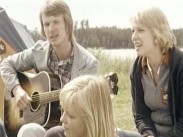 En ung man spelar akustisk gitarr omgiven av ett par kvinnor, i bakgrunden syns ett tält och en sjö.