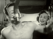En man utför ett konditionstest med en andningsventil fastspänd över ansiktet, leende kvinna i bakgrunden.