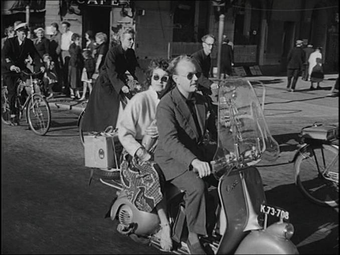 En man skjutsar en kvinna på en vespa bland flertalet cyklister och fotgängare i stadsmiljö.