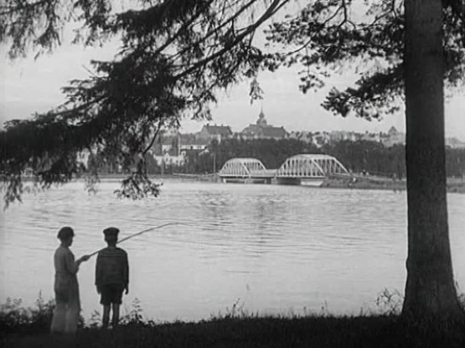 Ett par barn metar i förgrunden, bron till Frösön i bakgrunden så som den såg ut i slutet av 1920-talet.