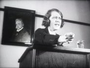 En kvinna med knuten näve och bestämd min i en talarstol, porträtt på Hjalmar Branting i bakgrunden.