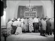 Ett femtontal vuxna knäböjer framför ett altarskåp, kvinnor till vänster och män till höger.
