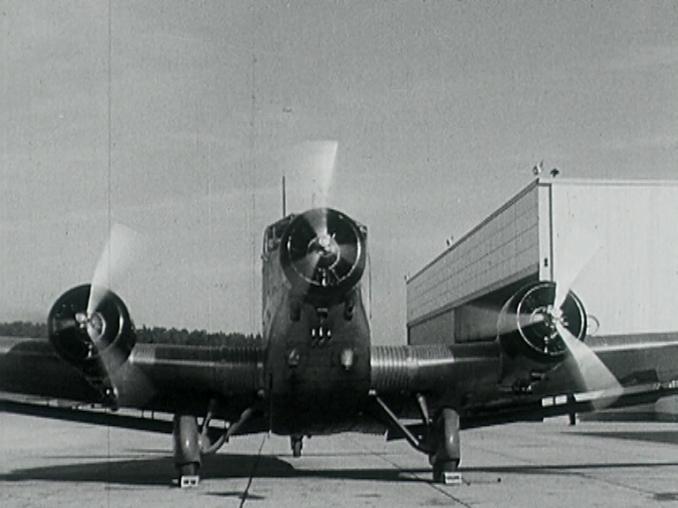 Ett propellerplan på marken fotat framifrån, flygplatsbyggnad till höger i bild.