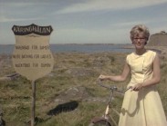 En kvinna med en cykel vid en skylt som lyder: KÄRINGHÅLAN - NAKENBAD FÖR DAMER, havet i bakgrunden.