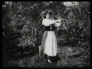 En kvinna i långkjol med förkläde och en krans i håret står i en trädgård.