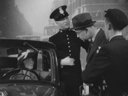 En kvinna sitter bakom ratten i en bil, utanför står en poliskonstapel, en man i hatt och en grabb i keps med cykel.