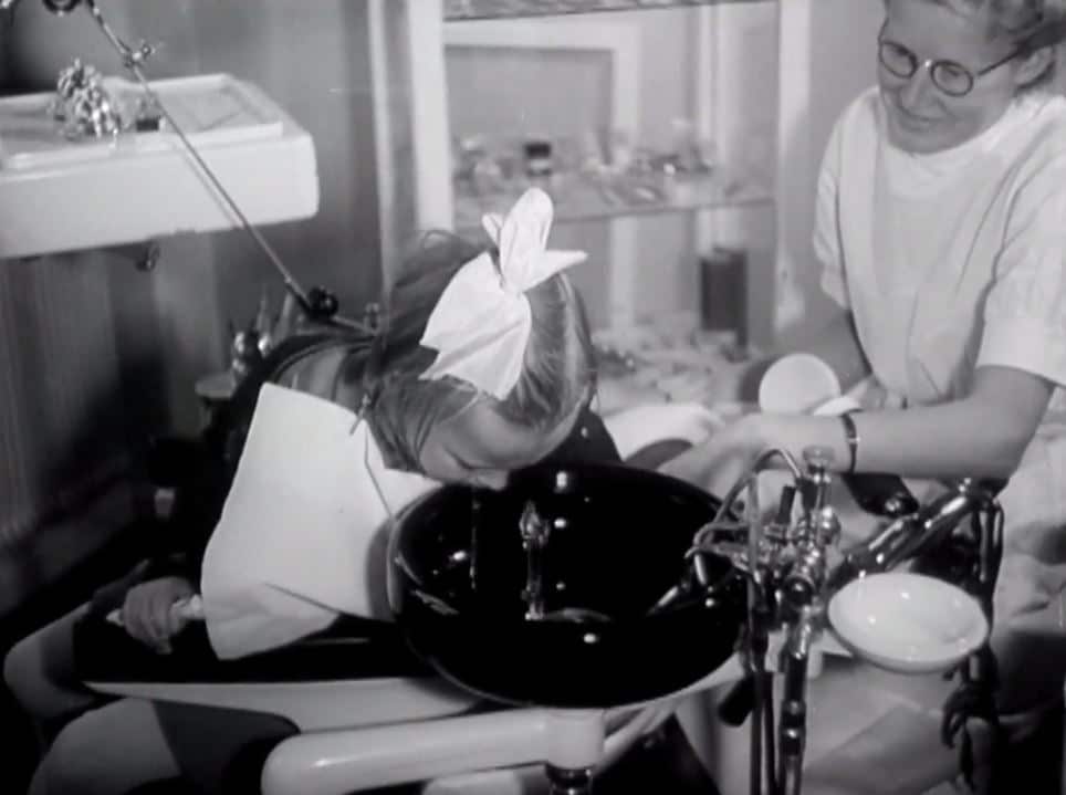 En liten flicka sitter i en tandläkarstol och sköljer munnen i en spottkopp, en sköterska sitter bredvid.