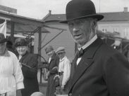 Man i hatt tittar in i kameran fotograferad på Stortorget i Växjö 1927, människor i bakgrunden också vända mot kameran.