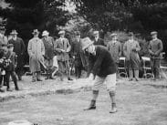 Golfare i hatt och knästrumpor vid utslagsplatsen, publik i bakgrunden.