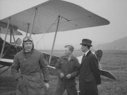 Man i flygarmössa med skyddsglasögon i pannan, mekaniker i overall samt en finklädd herre framför ett biplan.