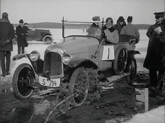 En förare av en öppen tävlingsbil sitter vid ratten omgiven av några människor, snö på marken.