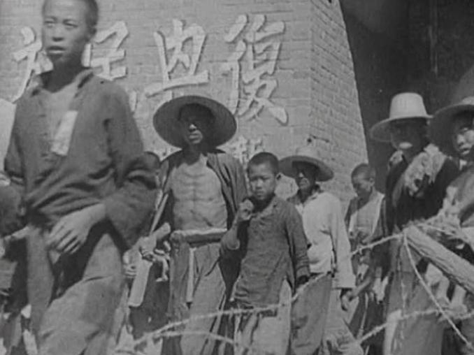En grupp människor på led, taggtråd i förgrunden och kinesiska tecken på en tegelvägg i bakgrunden.
