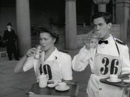 En servitris och en servitör med nummerlappar och varsin bricka i handen, båda dricker ur varsitt vattenglas.