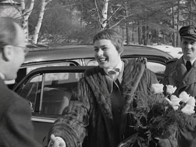 En leende Ingrid Bergman i päls och med blommor i famnen sträcker ut handen för att hälsa på Harald Molander, bil med chaufför i bakgrunden.