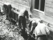 Fyra män gräver med spadar längs en husgrund på ett bygge.