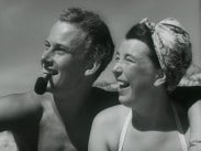 En man med pipa håller armen om en kvinna med sjalett, sanddyner i bakgrunden.