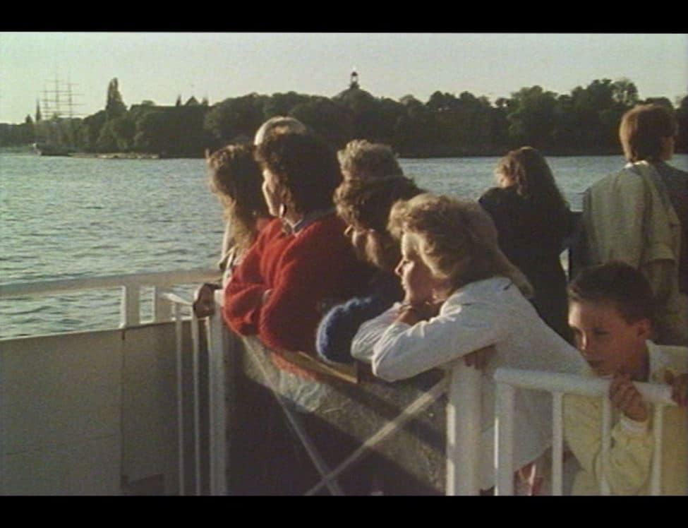 Ett tiotal passagerare utomhus på en färja i Stockholm, af Chapman syns i bakgrunden.