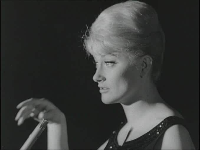 Monica Zetterlund i klänning framför en mikrofon, svart bakgrund.