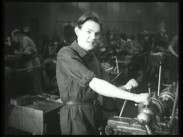 En ung man vid en maskin i fabriksmiljö, fler arbetare i bakgrunden.