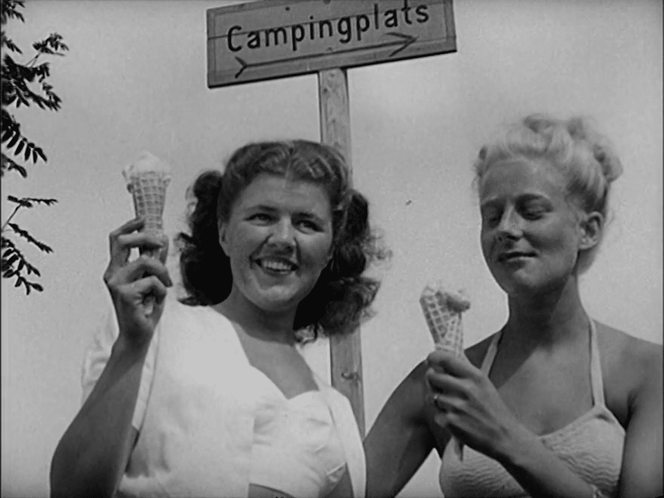 Två kvinnor äter glass, skylt med texten "Campingplats" i fonden.