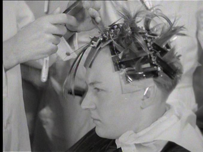Närbild av en ung man hos frisören som ska genomgå en kemisk permanent, hans hår är uppsatt med massor av klämmor.
