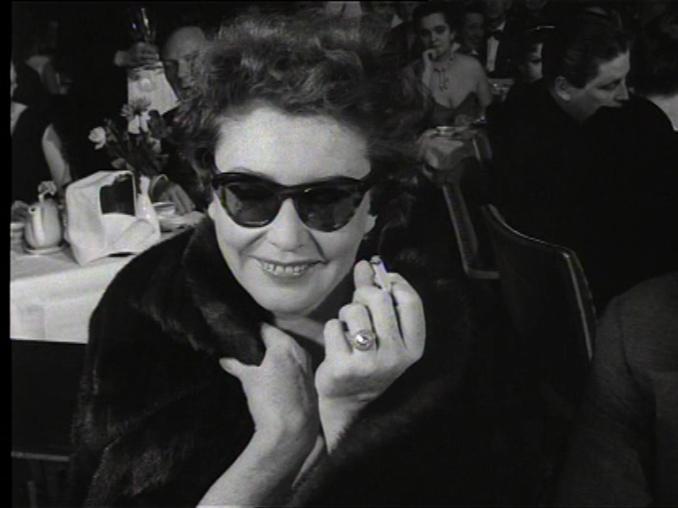 En leende Zarah Leander i mörka solglasögon och med en cigarett i handen sittandes på en restaurang, kroggäster i bakgrunden.