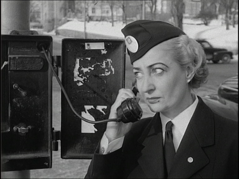 Uniformsklädd kvinna håller gammaldags telefonlur vid höger öra, bil i bakgrunden.