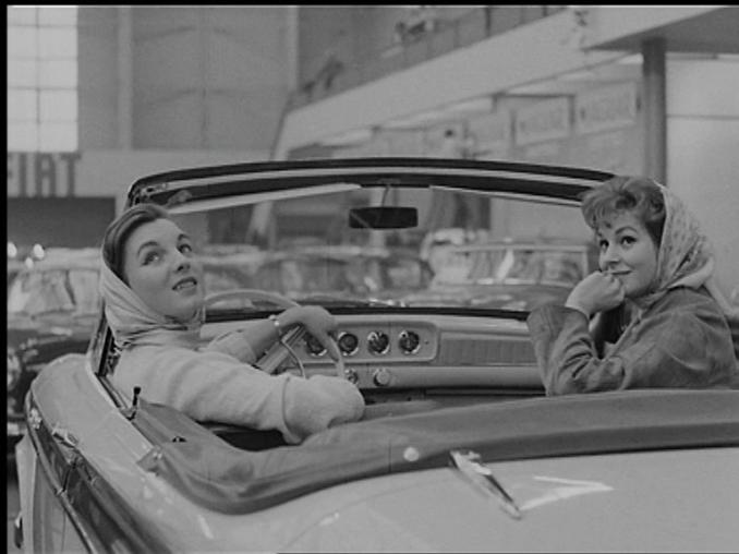 Två kvinnor i en Cabriolet på en bilutställning inomhus, fler bilar och en FIAT-skylt i bakgrunden.
