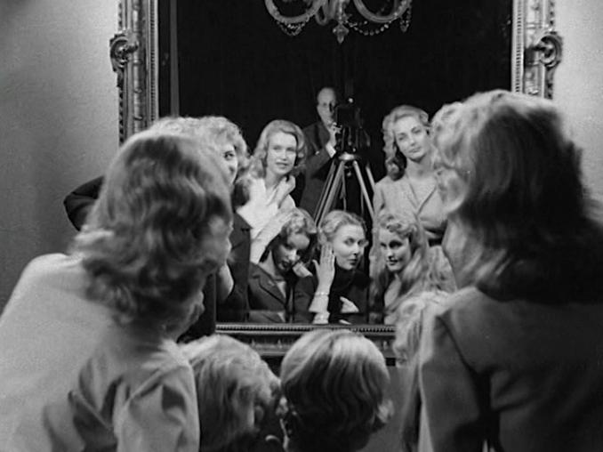 En handfull unga kvinnor som ordnar sitt hår samt en filmfotograf framför en stor spegel.