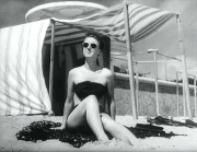 En kvinna i baddräkt och solglasögon på en sandstrand, omklädningshytt i bakgrunden.