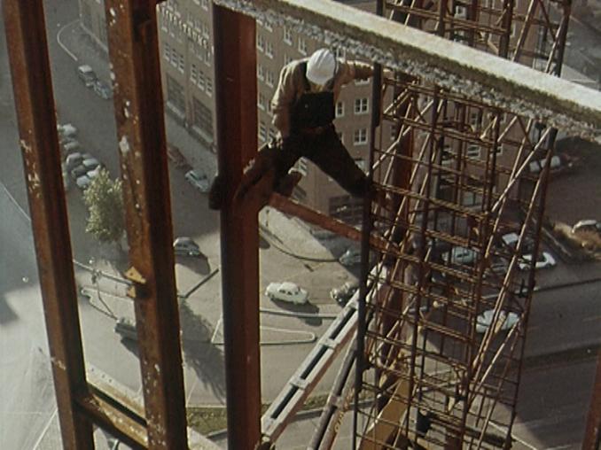 En byggnadsarbetare högt ovanför marken bland bjälkar och betong under uppförandet av ett högt hus.