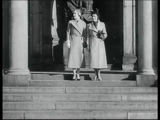 Två kvinnor i kappor på en trappa framför en byggnad.