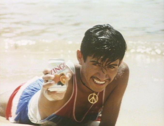En man ligger på mage på en strand i vattenbrynet och håller upp en läskburk av märket Zingo.